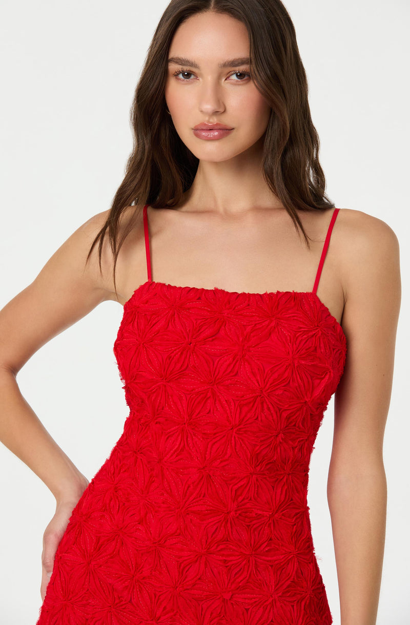 A woman is wearing a sleeveless red dress with thin spaghetti straps. The dress features an intricate textured floral pattern throughout.