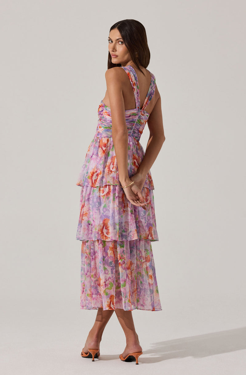 A woman is wearing a sleeveless, tiered dress with a floral pattern in shades of pink, purple, and orange. The dress has wide straps and a fitted bodice with a slight V-neckline. The skirt consists of three layers of flowing, semi-sheer fabric with a colorful floral design. She is also wearing open-toe high heel shoes and a delicate bracelet on her left wrist.