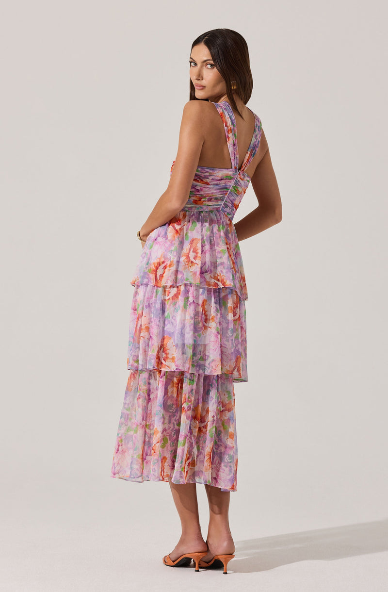 A woman is wearing a sleeveless, knee-length dress with wide shoulder straps. The dress features a colorful, floral pattern with shades of pink, purple, orange, and green. The top of the dress has a smocked bodice with gathered fabric, and the skirt consists of multiple tiered, ruffled layers.