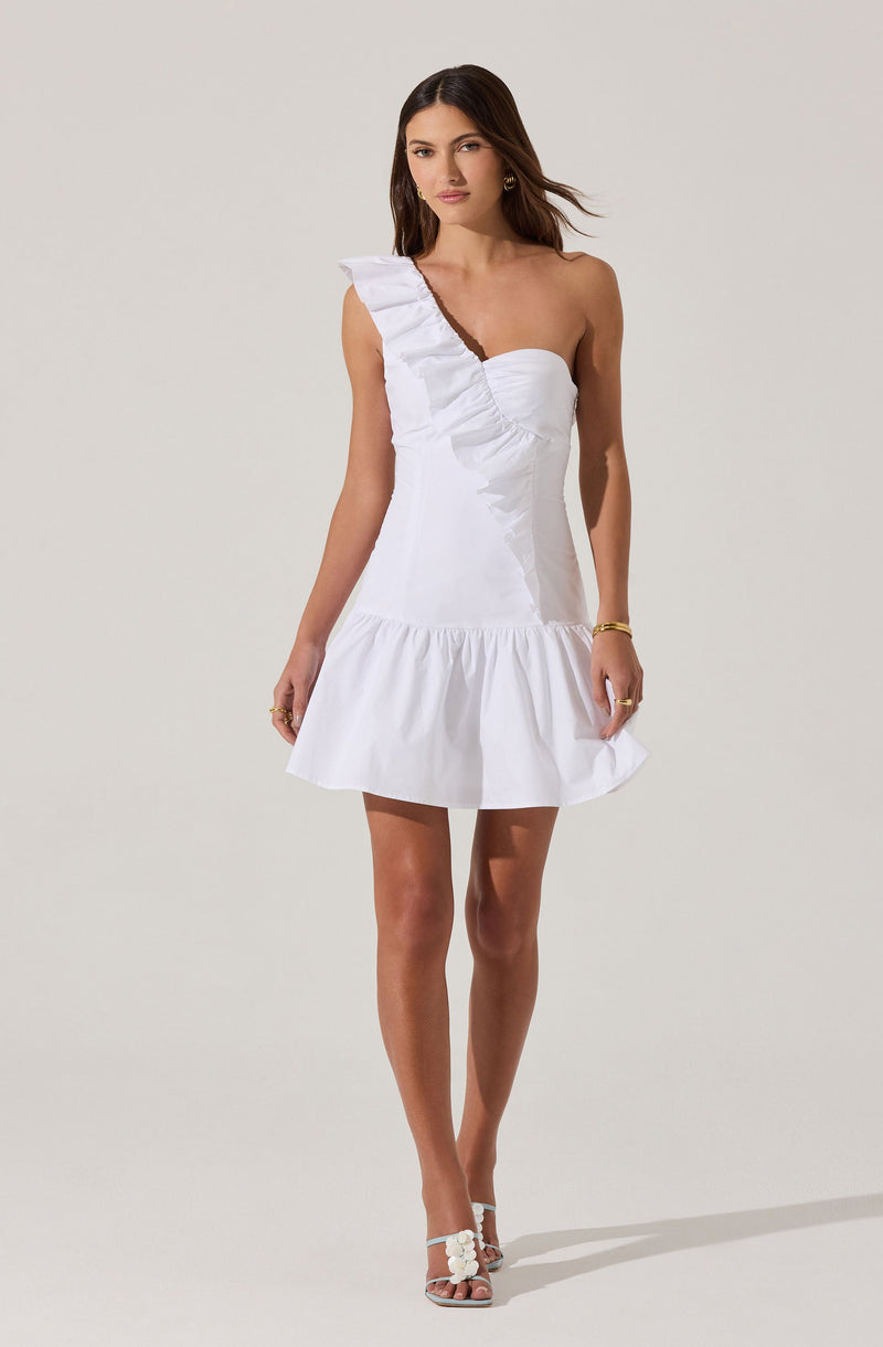 A woman is wearing a short, white, sleeveless dress with an asymmetrical one-shoulder design. The dress features ruffle detailing along the shoulder strap and a gathered, flared hem at the bottom. She is also wearing open-toe high heels with flower embellishments.