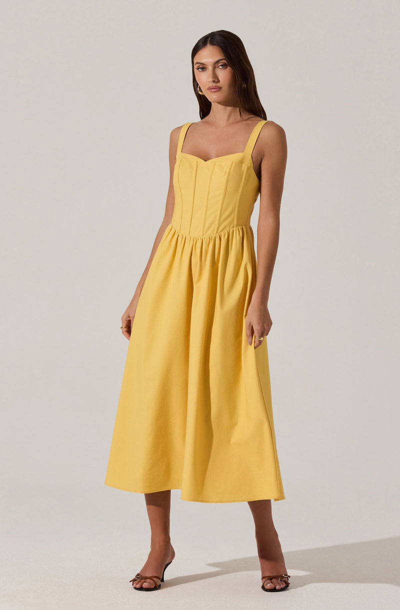 A woman is wearing a sleeveless, yellow sundress with wide shoulder straps and a fitted bodice that features vertical seam details. The dress has a gathered waistband and a full, flowing skirt that falls to mid-calf length. She is also wearing open-toed heels and minimal jewelry.