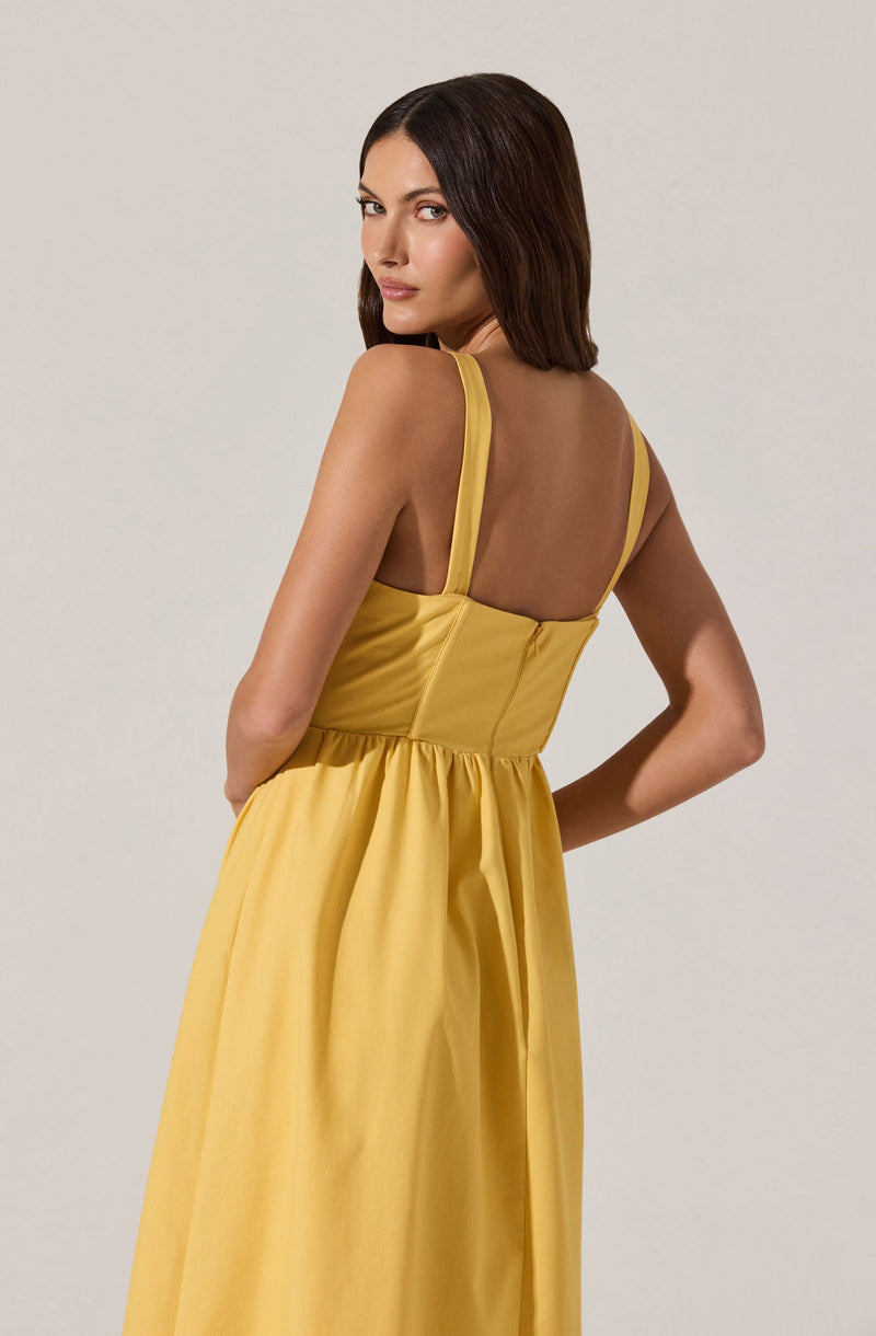 A woman is wearing a yellow dress with wide shoulder straps and a fitted bodice. The back of the dress features a sheer panel that covers the upper back, adding a delicate and stylish detail. The skirt of the dress is long and gathered at the waist, creating a flowing silhouette.