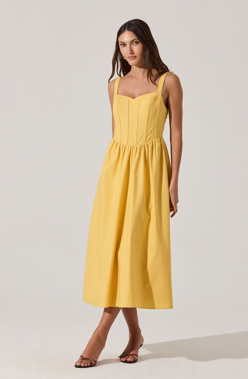 A woman is wearing a sleeveless, yellow dress with wide shoulder straps and a fitted bodice that features vertical seam details. The dress has a gathered waistline and a full, midi-length skirt that falls to mid-calf. She pairs the dress with open-toe, high-heeled sandals.