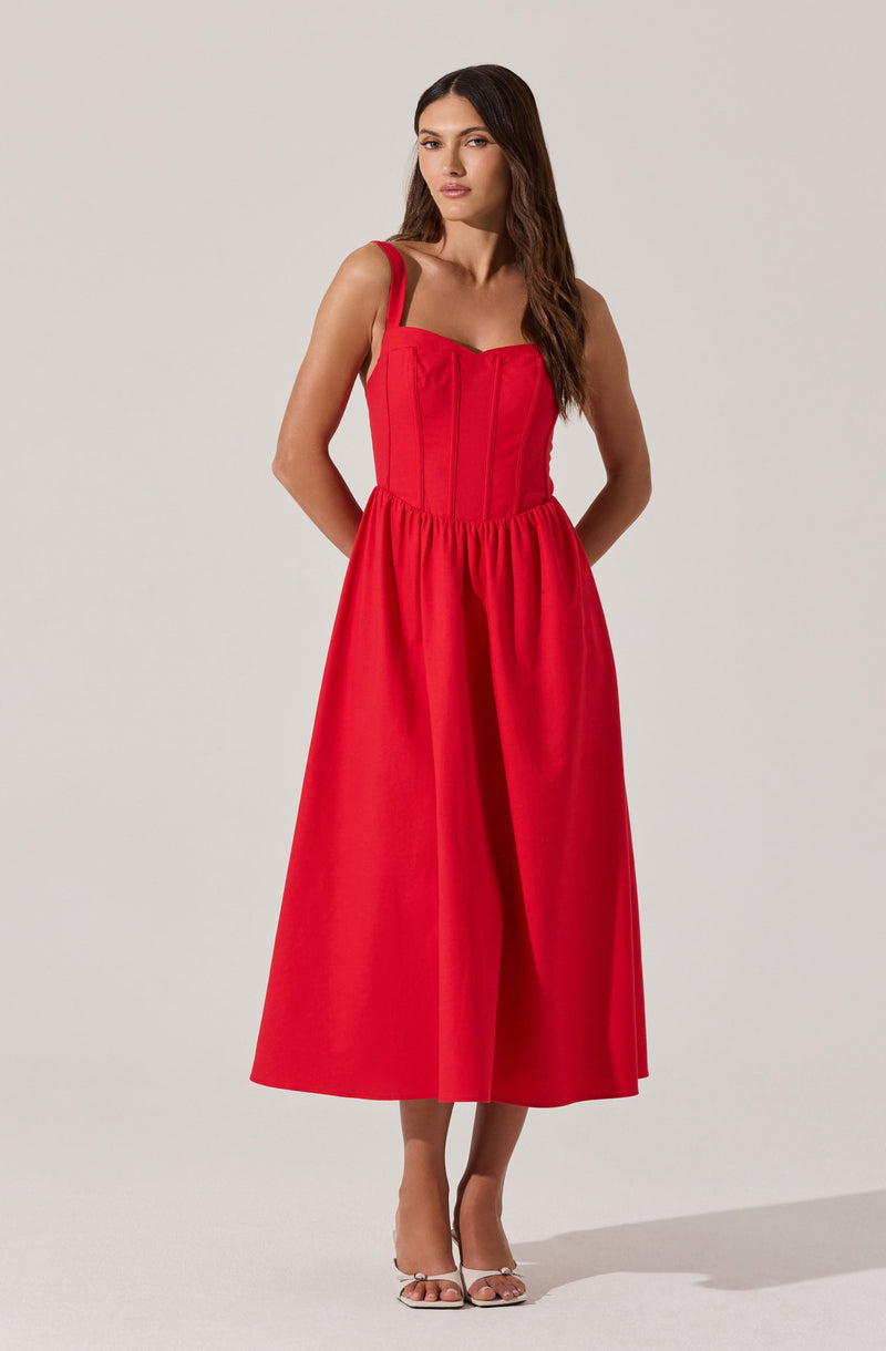 A woman is wearing a sleeveless, bright red dress with wide shoulder straps. The dress has a fitted bodice with vertical seam details and a gathered, full-length skirt that reaches mid-calf. She is also wearing white high-heeled sandals.