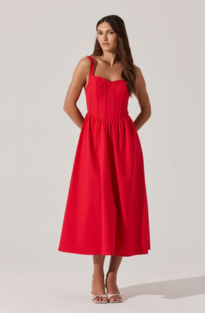 A woman is wearing a sleeveless, bright red dress with wide shoulder straps. The dress has a fitted bodice with vertical seam details and a gathered, full-length skirt that reaches mid-calf. She is also wearing white high-heeled sandals.