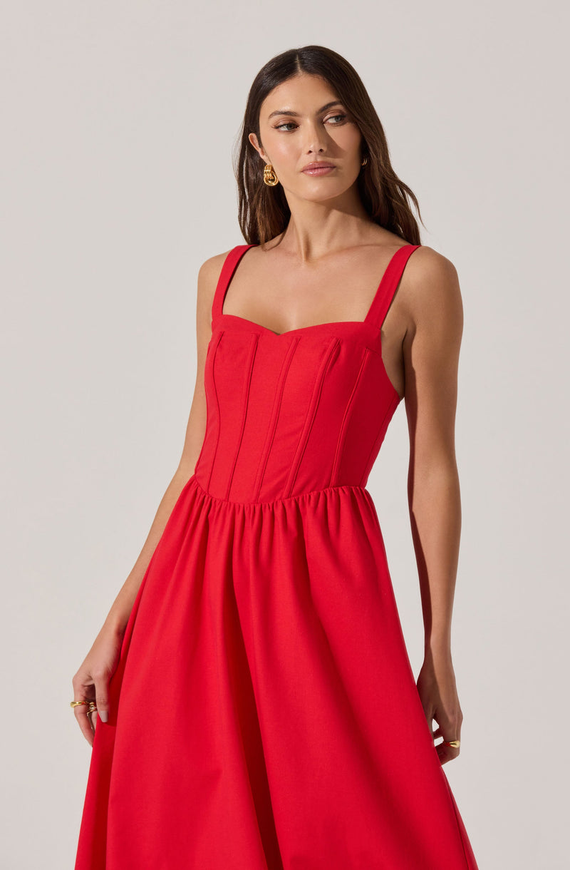 A woman is wearing a sleeveless red dress with wide shoulder straps. The bodice of the dress features vertical seam details, and the skirt is gathered at the waist, creating a full, flowing silhouette.
