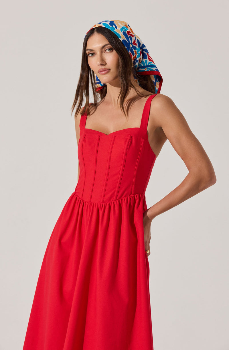 A woman is wearing a sleeveless, bright red dress with a fitted bodice that features vertical seams and a sweetheart neckline. The skirt is full and gathered at the waist. She has a colorful scarf with a floral pattern in shades of blue, red, beige, and white wrapped around her head.
