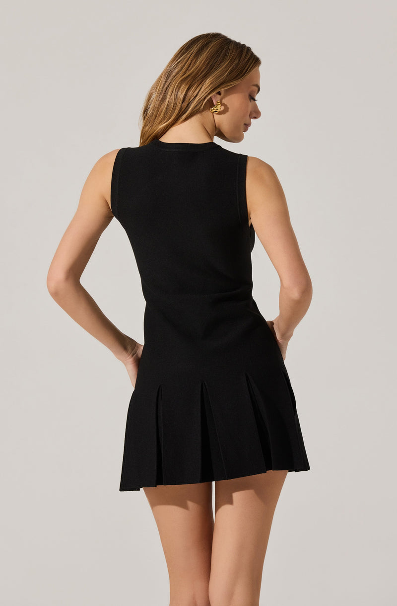 A woman is wearing a sleeveless black dress with a fitted bodice and a pleated skirt. The dress has a round neckline and the pleats on the skirt create a structured, stylish look. She is accessorized with gold earrings and has her hair styled in loose waves.