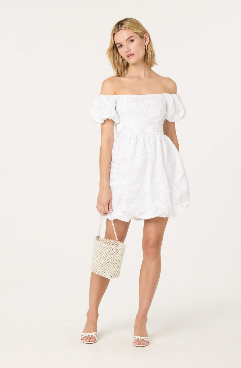 A woman is wearing an off-the-shoulder white dress with puffed short sleeves and a gathered waist. She is holding a small, beaded handbag with a chain strap and is wearing open-toed white high heels.