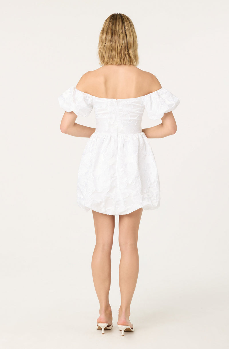 A woman is wearing a white off-the-shoulder dress with puffed short sleeves and a fitted bodice. The dress has a textured, slightly floral pattern and ends above the knees. She is also wearing white high-heeled shoes.