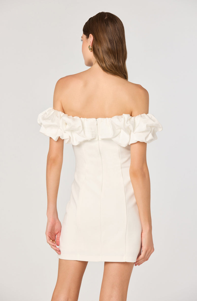 A woman is wearing an off-the-shoulder dress with ruffled sleeves. The dress is fitted, in a light cream or beige color, and has vertical seams running down the back. She is accessorized with gold earrings.