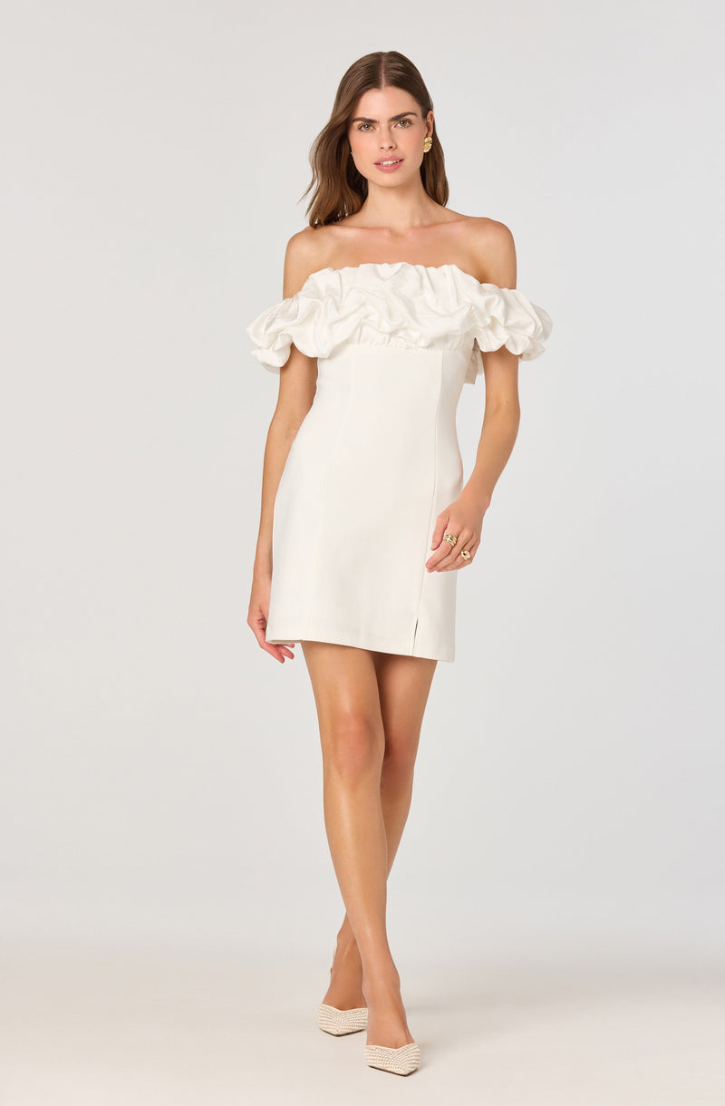 A woman is wearing an off-the-shoulder white dress with a ruffled neckline detail. The dress is fitted and short in length, ending mid-thigh. She is also wearing beige high-heeled shoes.