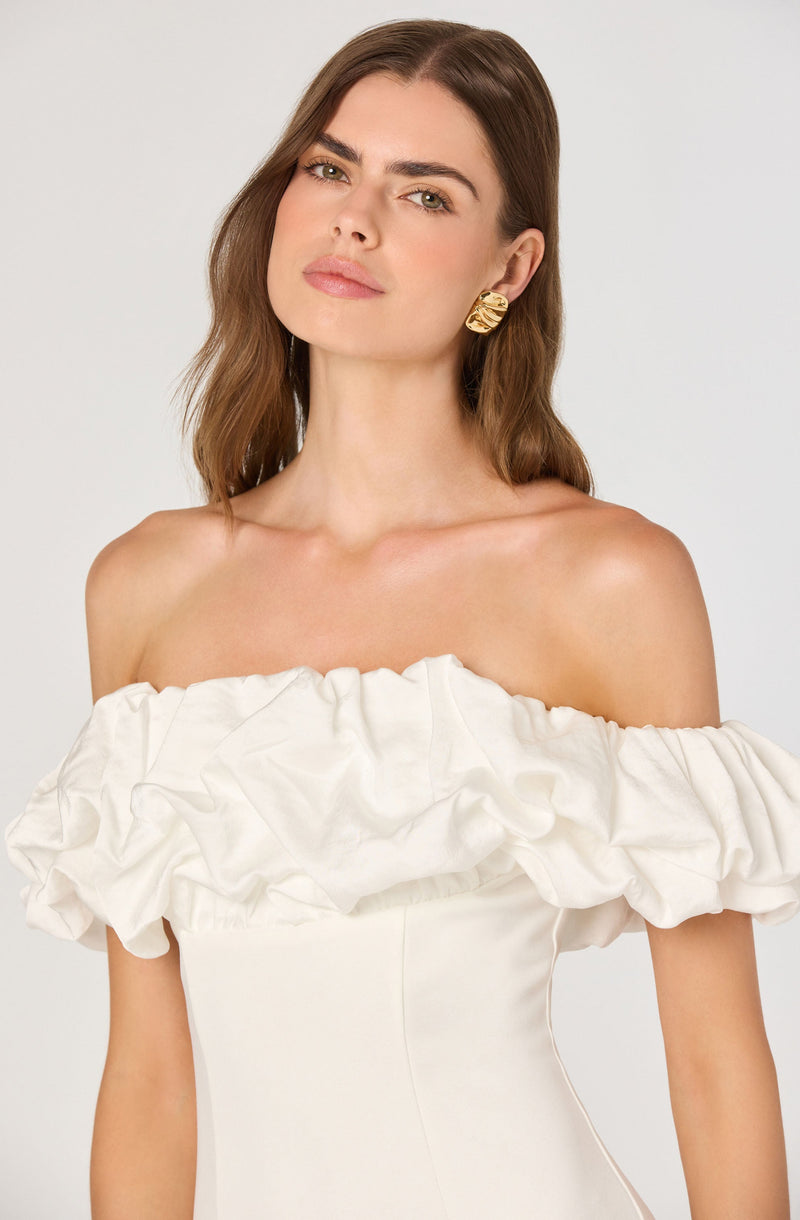 A woman is wearing an off-the-shoulder white dress with a large, ruffled, and gathered fabric detail around the neckline. She has on bold gold earrings and has shoulder-length brown hair.