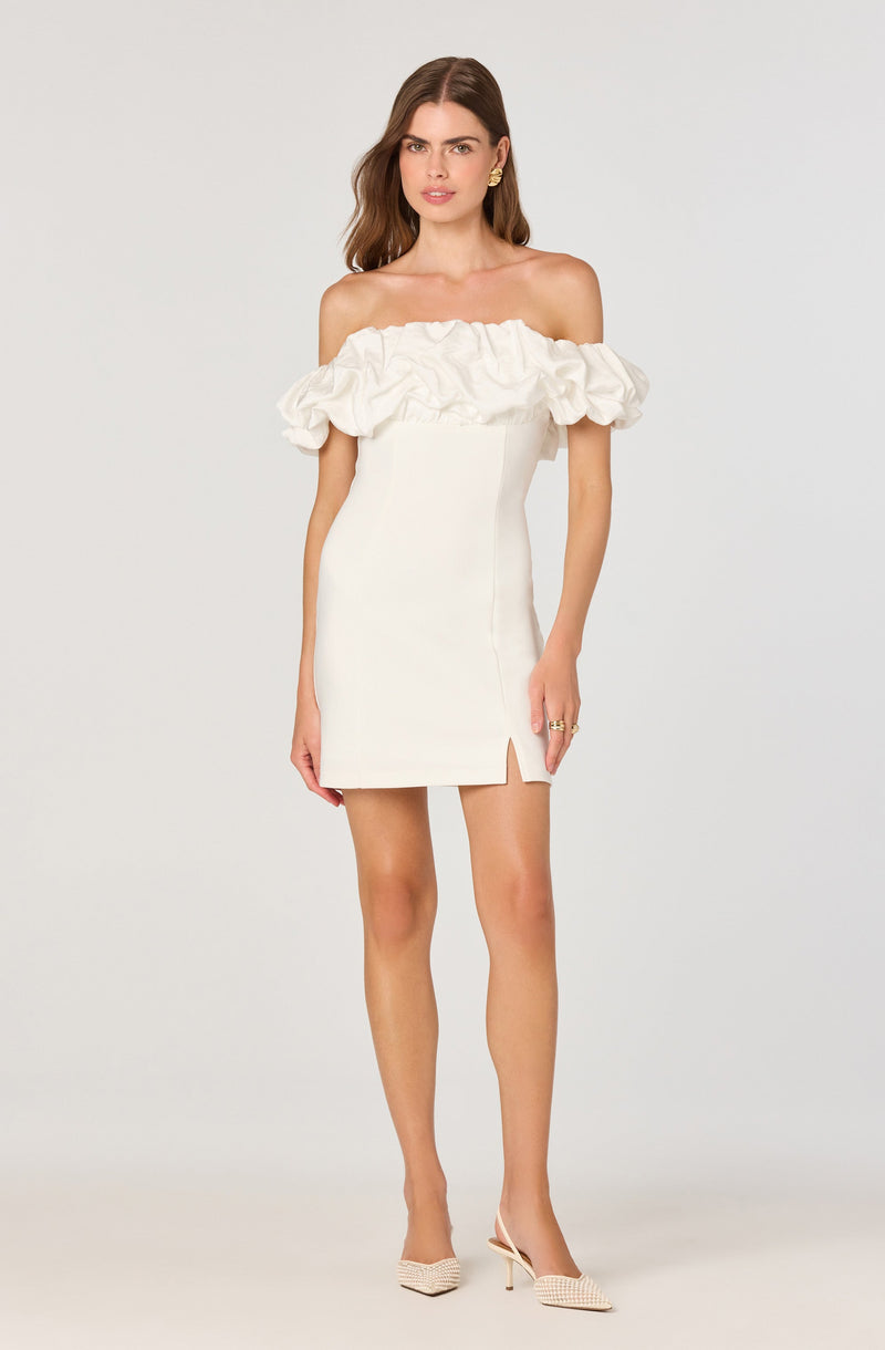 The dress is an off-the-shoulder style with a ruffled neckline and a fitted, straight silhouette. It is white in color and features a small slit on the front left side. The model is wearing beige high-heeled shoes with a textured pattern.
