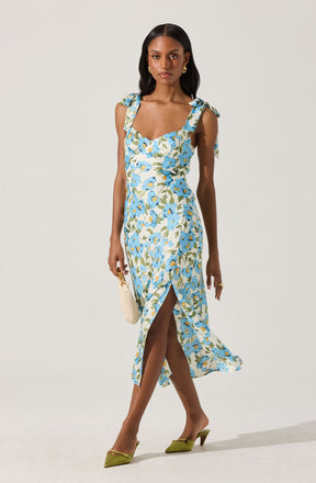 The woman is wearing a sleeveless, ankle-length dress with a light-colored background and a floral pattern featuring blue, white, and green colors. The dress has shoulder straps tied into bows. She is also wearing green pointed-toe high heels and carrying a small, white handbag.
