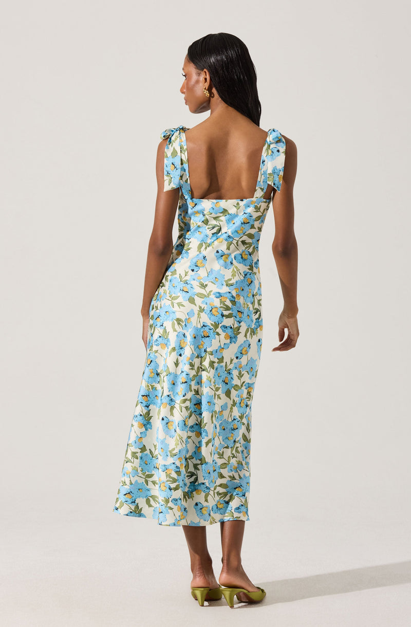 A woman is wearing a long, sleeveless dress with shoulder tie straps. The dress features a light background with a blue floral pattern and green leaves. She is wearing green high-heeled shoes.