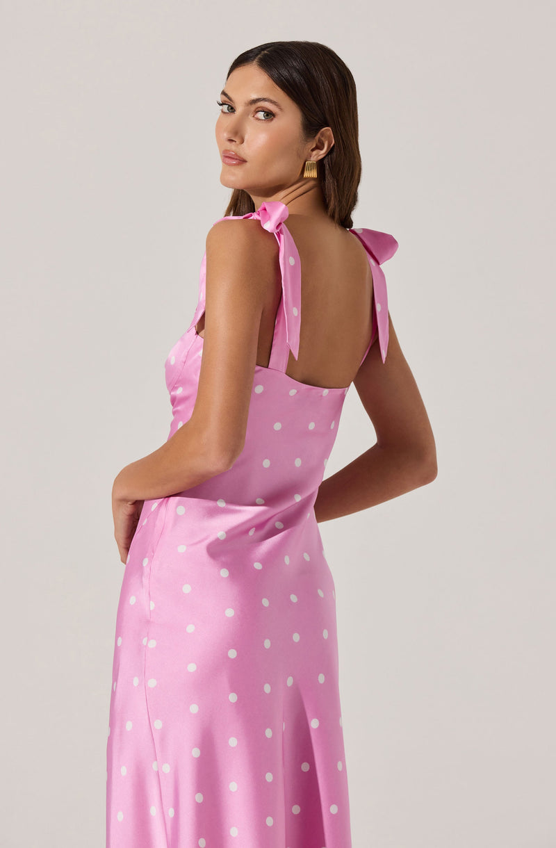 A woman is wearing a pink sleeveless dress with white polka dots. The dress has shoulder straps tied into bows on each shoulder.