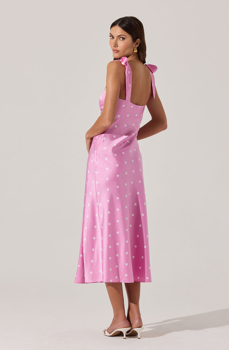 A woman is wearing a pink sleeveless midi dress with white polka dots. The dress features shoulder straps tied into bows on each shoulder and a fitted bodice that flows into a slightly flared skirt. She is also wearing white heeled sandals.