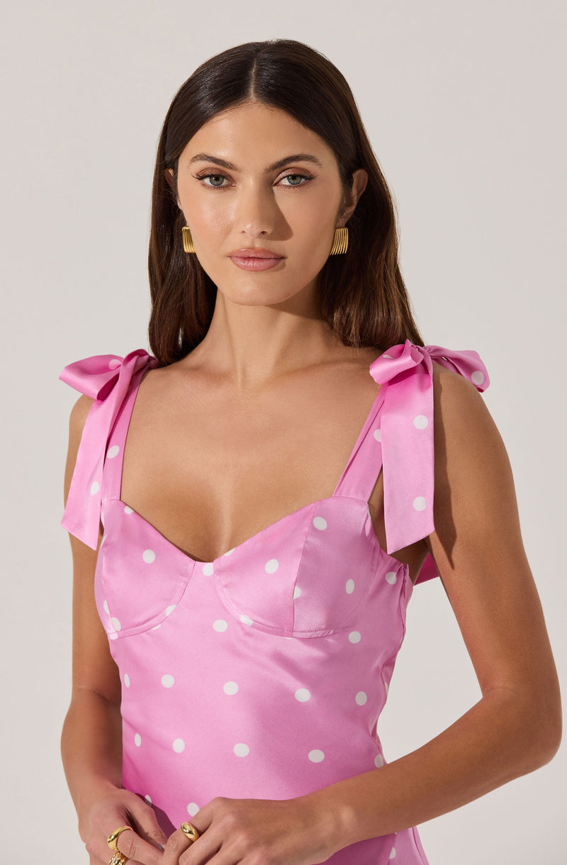 A woman is wearing a pink dress with white polka dots. The dress features shoulder straps tied into bows and has a fitted bodice with visible seam details. She accessorizes with gold earrings and rings.