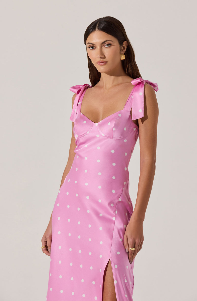 A woman is wearing a pink satin dress with white polka dots. The dress features shoulder straps tied into bows, a fitted bodice, and a thigh-high slit on the side.