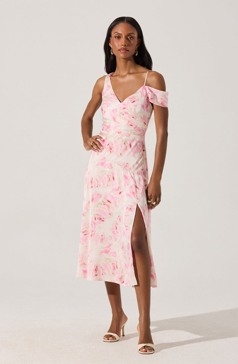 A woman is wearing a light pink and white tie-dye midi dress with thin spaghetti straps, an off-shoulder ruffle detail on one side, and a high slit on the left side. She is also wearing beige open-toe high heels and gold jewelry.