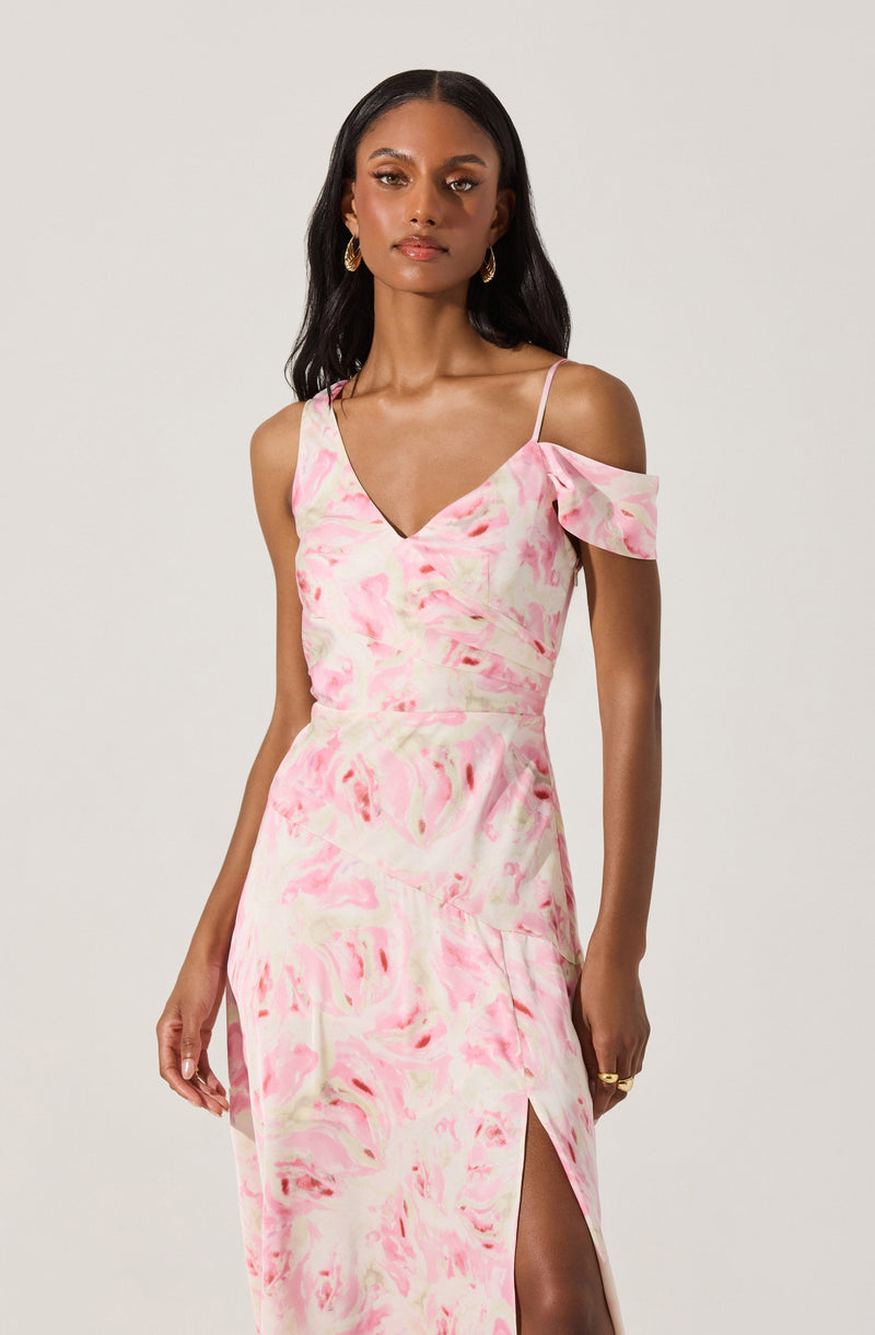 A woman is wearing a pastel pink and cream tie-dye dress with a V-neckline and thin spaghetti straps. The dress features a cold-shoulder ruffle detail on one side and a high slit on the leg.