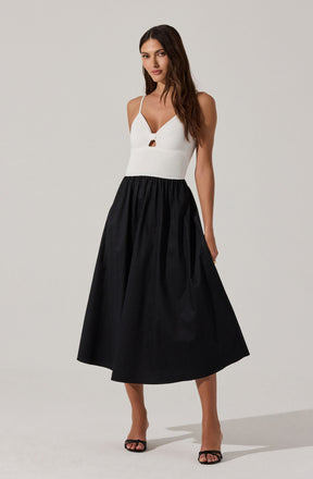 A woman is wearing a sleeveless, white, ribbed knit top with thin spaghetti straps and a small keyhole cutout at the front. The top is fitted at the waist. She pairs it with a high-waisted, full-length black skirt that has a gathered, voluminous silhouette and reaches just above her ankles. She is also wearing black open-toe heels.