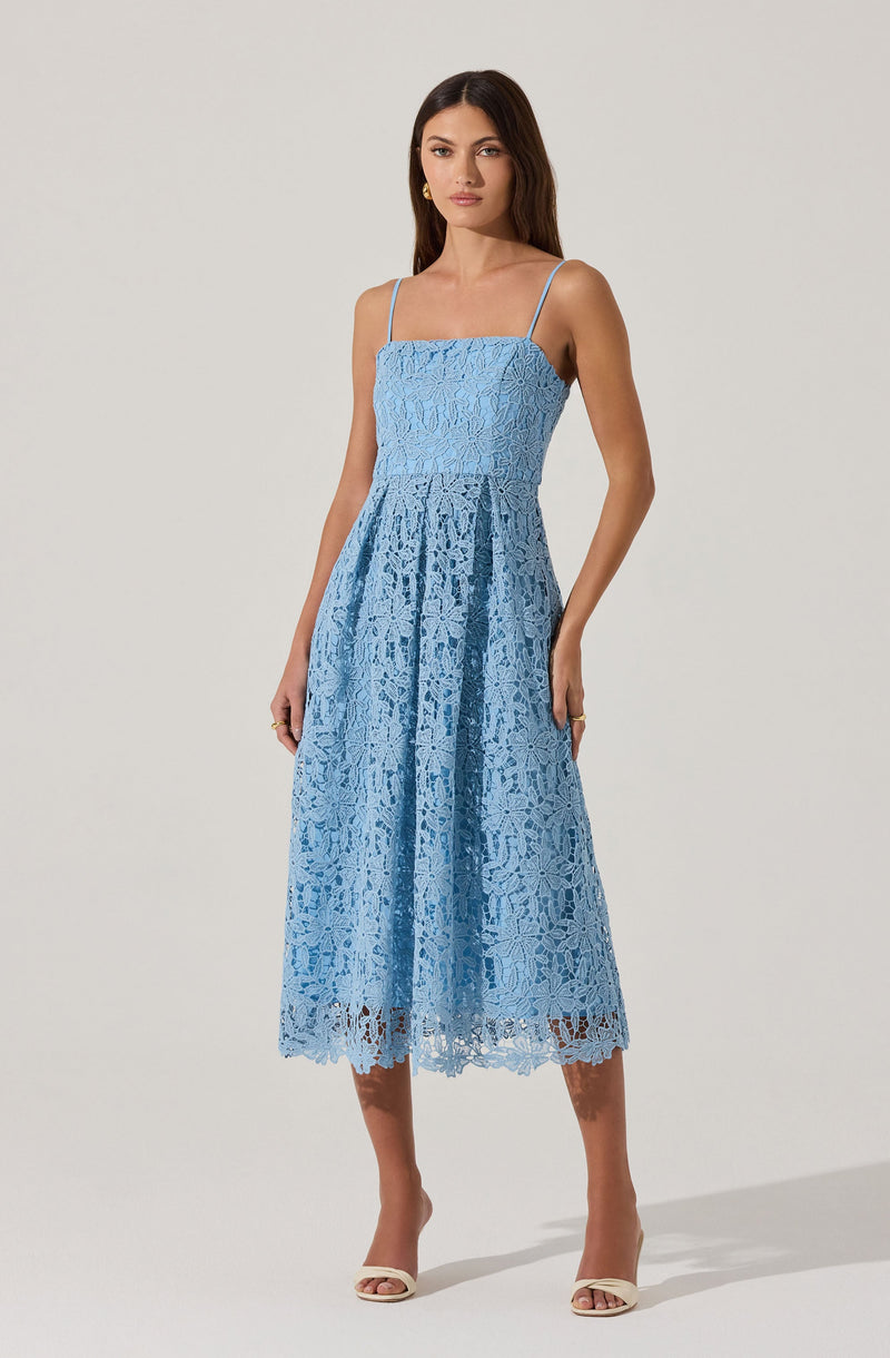 A woman is wearing a light blue, sleeveless dress with thin spaghetti straps. The dress features a fitted bodice and a skirt with a lace overlay that extends to mid-calf length, with a scalloped hemline. The lace pattern is floral and intricate throughout the dress. She is also wearing open-toe heels and gold hoop earrings.