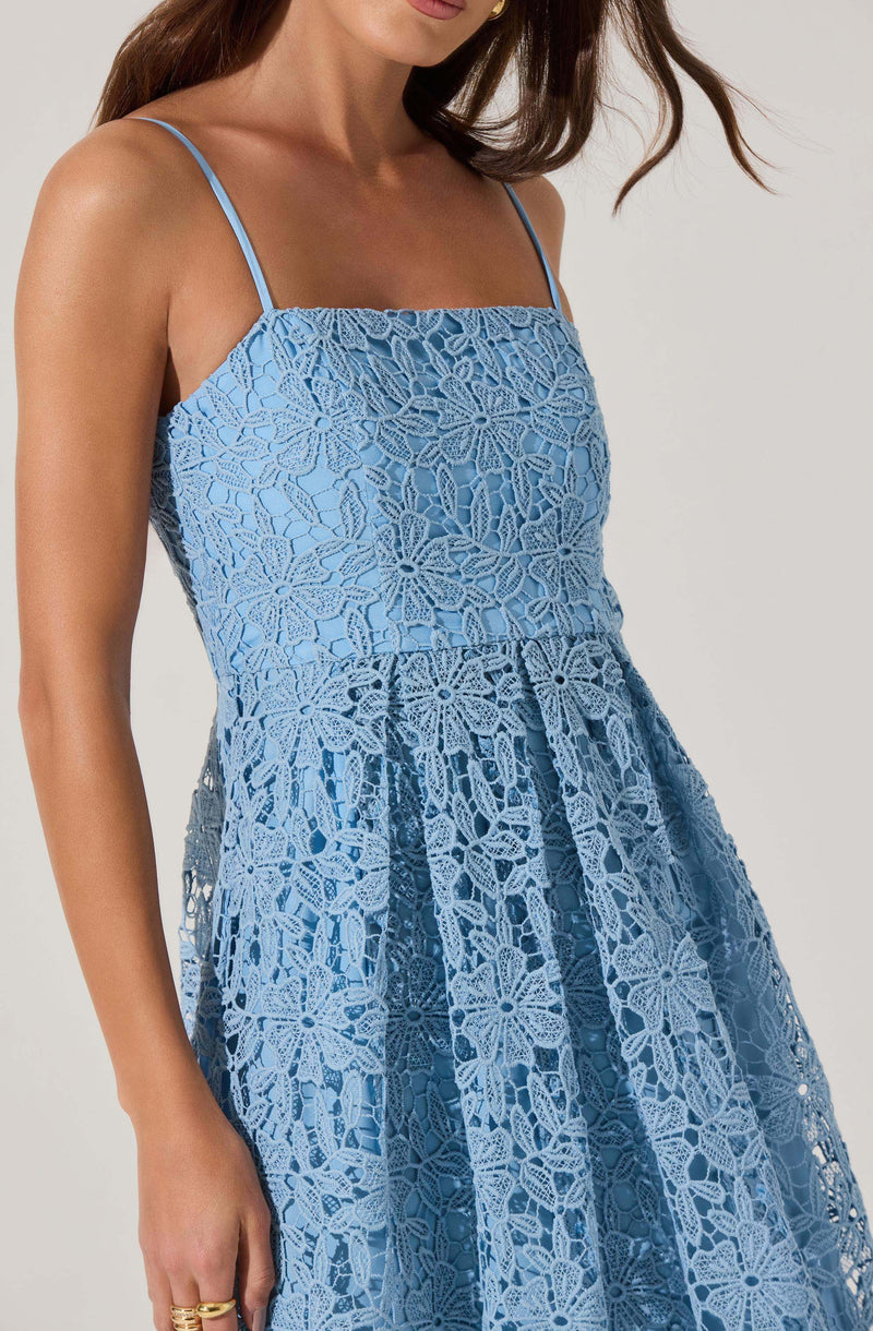 A woman is wearing a light blue dress with thin spaghetti straps. The dress features an intricate lace overlay with floral and geometric patterns, creating a textured and delicate appearance. The skirt of the dress has pleats, adding volume and movement.