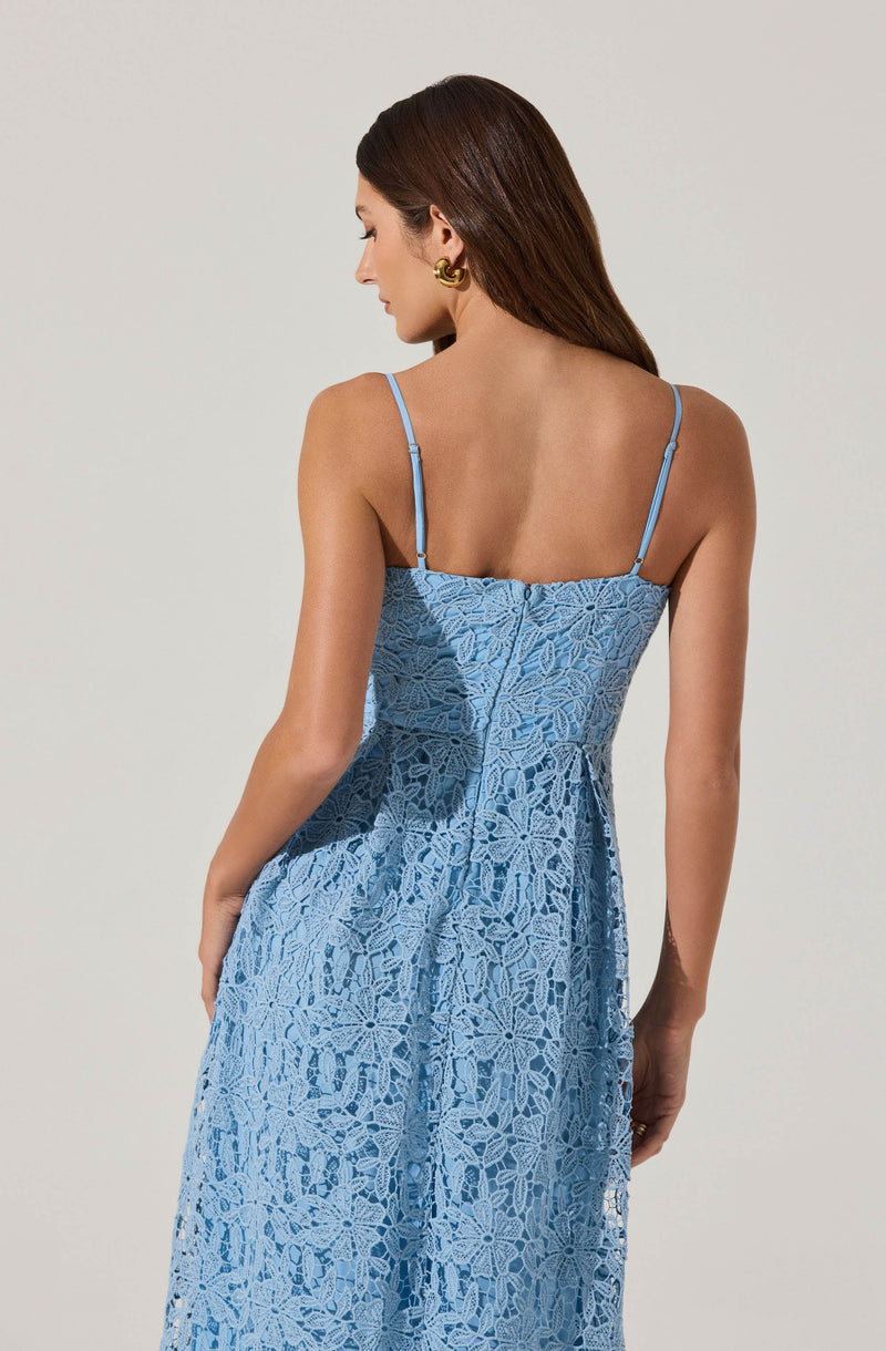A woman is wearing a light blue, sleeveless dress with thin spaghetti straps. The dress features an intricate lace pattern throughout, creating a textured and elegant appearance. The back of the dress is fitted at the waist and flows down in a loose, flowing style. The woman has long, dark brown hair and is accessorizing with a large, gold statement earring.