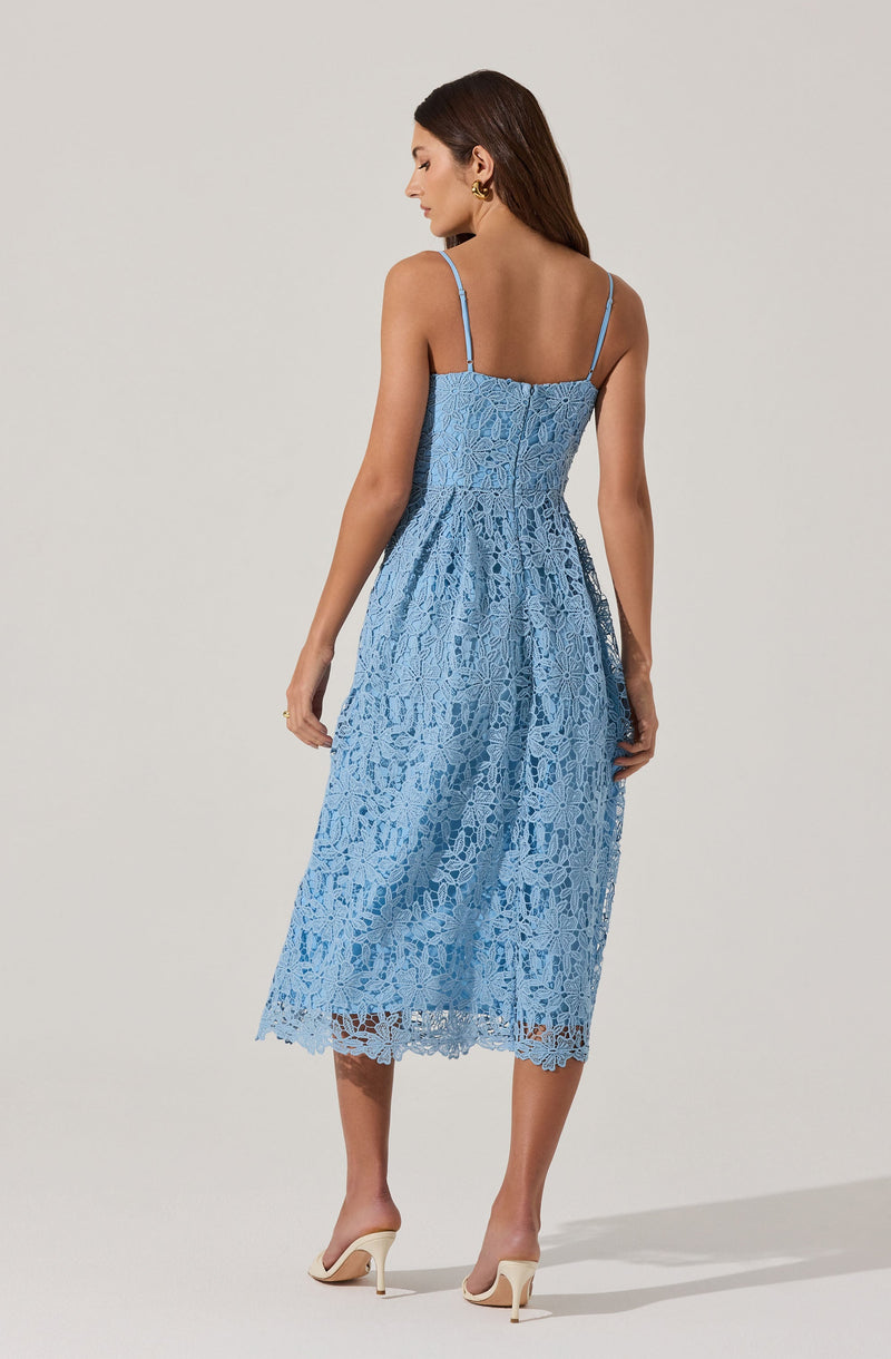 A woman is wearing a light blue lace midi dress with thin spaghetti straps. The dress features an intricate floral lace pattern and has a fitted bodice that flows into a slightly gathered skirt. She pairs the dress with cream-colored high heels.