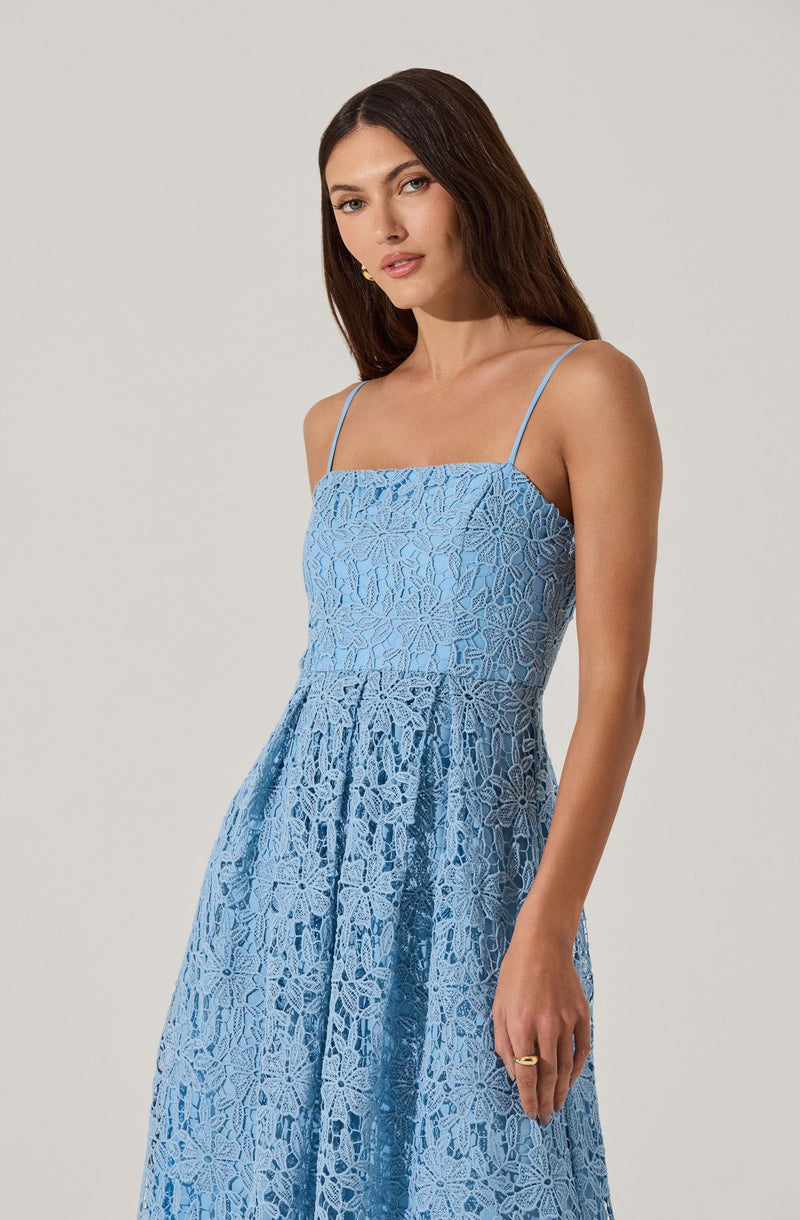 A woman is wearing a light blue dress made of lace fabric with a floral pattern. The dress features thin spaghetti straps and a fitted bodice that flows into a gathered skirt. The lace material is intricate and see-through in some areas, creating a delicate and elegant appearance.