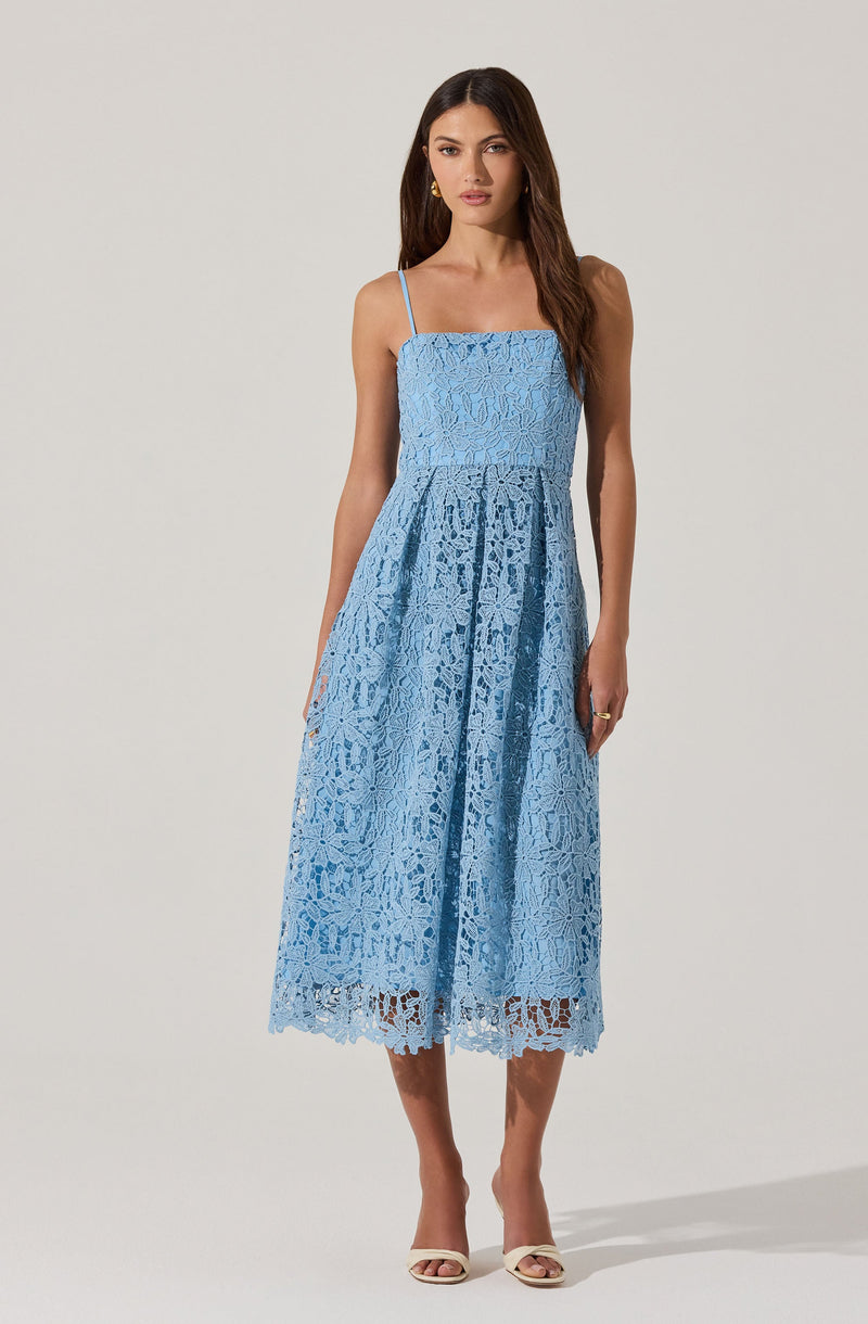 A woman is wearing a light blue lace dress with spaghetti straps. The dress has a fitted bodice, a high waistline, and a full-length skirt that features a scalloped hem. The lace fabric has a floral pattern throughout. She is also wearing jewelry, including earrings and rings, and has on white high-heeled sandals.