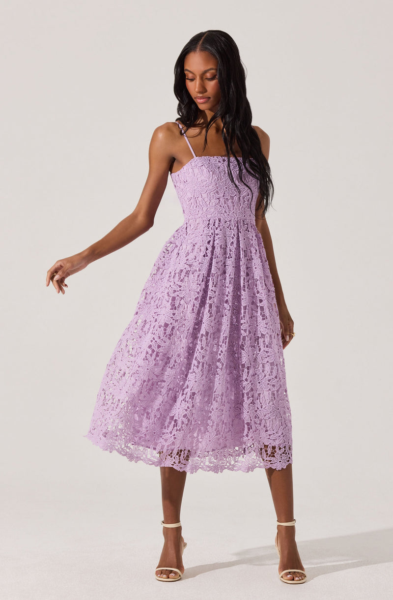 A woman is wearing a sleeveless, lavender-colored dress with thin spaghetti straps. The dress features a fitted, smocked bodice and a full, midi-length skirt made of lace fabric with an intricate floral pattern. She is also wearing nude high-heeled sandals.