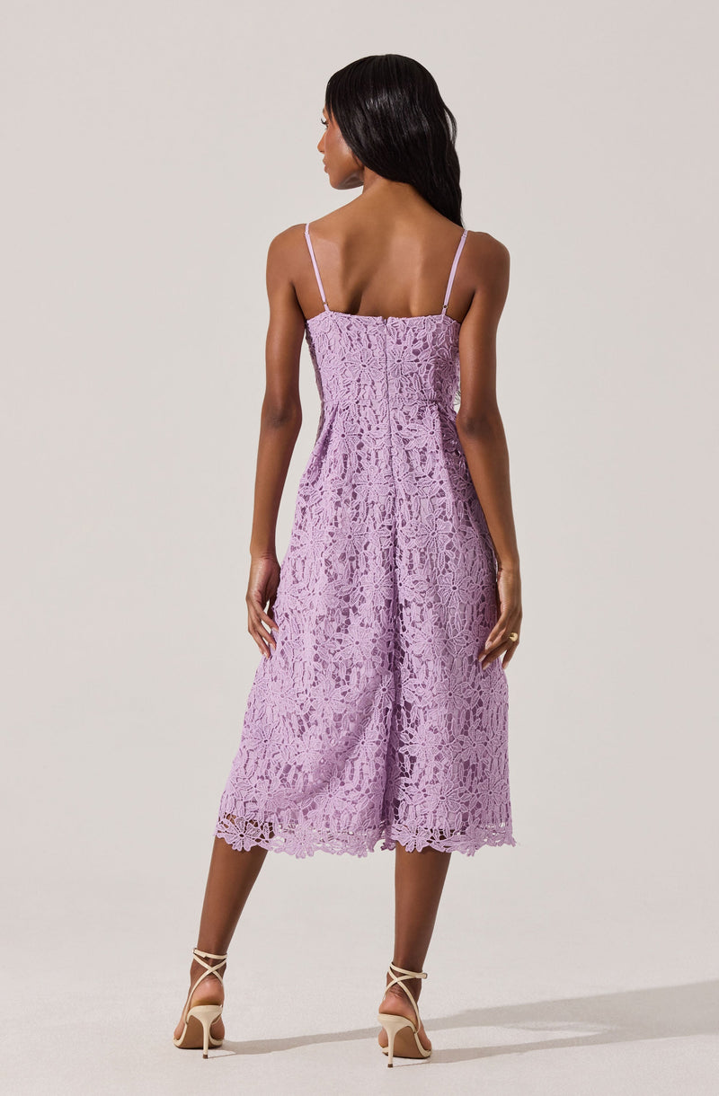 A woman is wearing a lavender-colored, sleeveless lace dress with thin spaghetti straps. The dress has a fitted bodice and a loose, midi-length skirt with a scalloped hem. She is also wearing strappy high-heeled sandals.