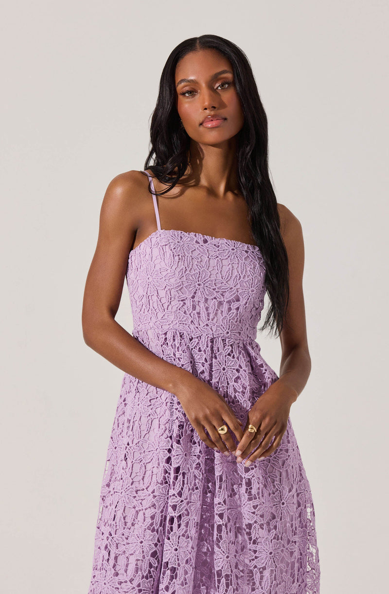 A woman is wearing a sleeveless, lavender-colored dress with thin spaghetti straps. The dress features an intricate lace pattern throughout, giving it a textured and floral appearance.
