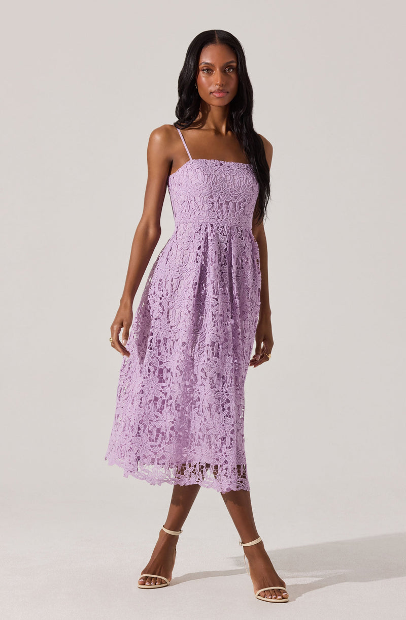A woman is wearing a light purple lace dress with thin spaghetti straps, a fitted bodice, and a midi-length skirt with a scalloped hem. She is also wearing nude high-heeled sandals.