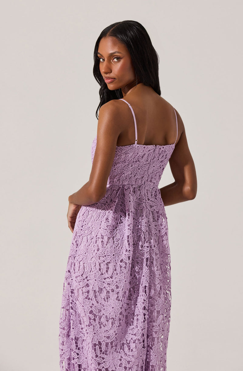 A woman is wearing a sleeveless, lavender-colored dress made of intricate lace fabric. The dress has thin spaghetti straps and a fitted bodice, flowing into a long, wide skirt that extends to the floor. The lace pattern features floral and geometric designs, creating a delicate and elegant look.