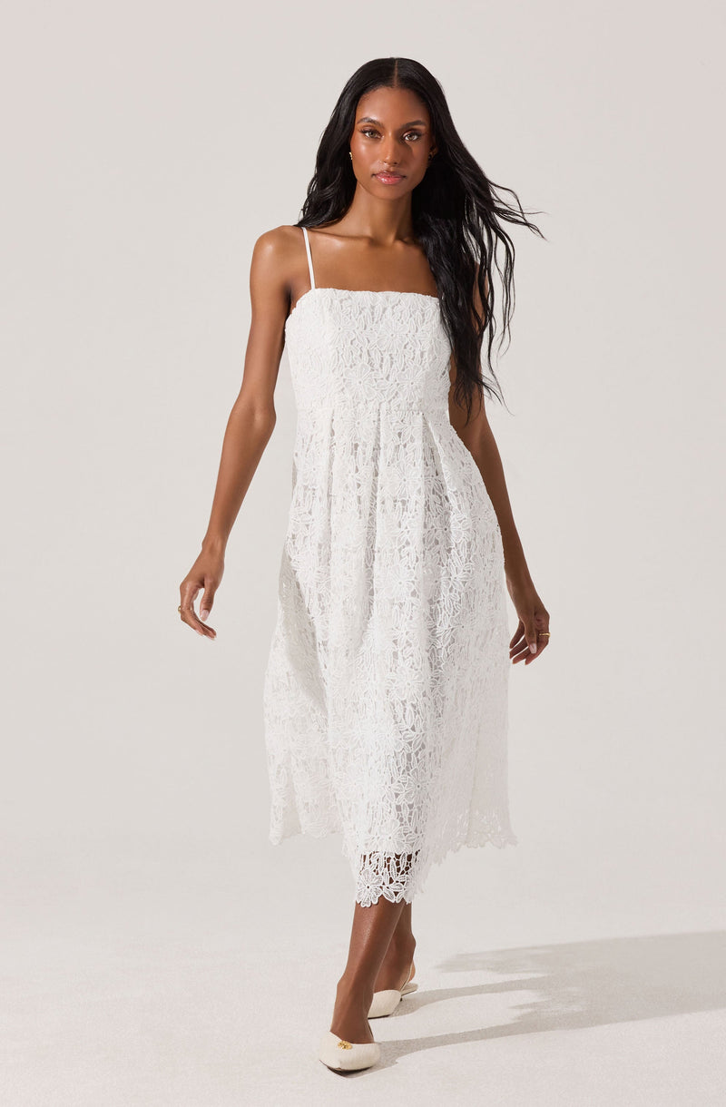 A woman is wearing a white, sleeveless dress with thin spaghetti straps. The dress features a textured lace pattern, a fitted bodice, and a flowy, mid-length skirt with a slightly scalloped hem. She is also wearing cream-colored shoes with small bows on the front.