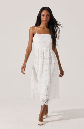 A woman is wearing a white, sleeveless dress with thin spaghetti straps. The dress features a textured lace pattern, a fitted bodice, and a flowy, mid-length skirt with a slightly scalloped hem. She is also wearing cream-colored shoes with small bows on the front.