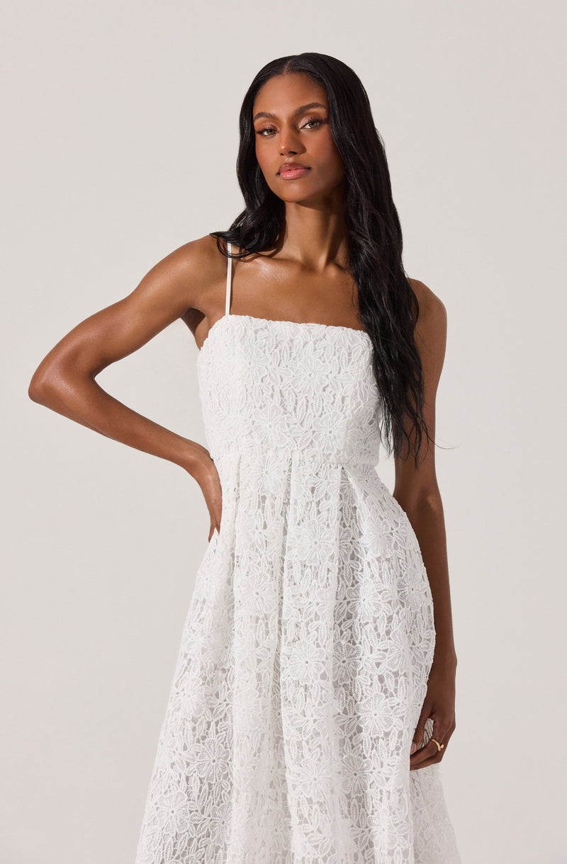 A woman is wearing a white sleeveless dress with thin spaghetti straps. The dress features an intricate lace pattern throughout, giving it a delicate and textured appearance. The fabric is semi-sheer with a floral lace design. The dress has a fitted bodice and a flowing skirt.