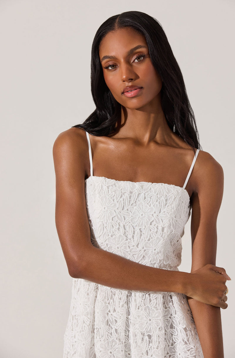 A woman is wearing a white dress with a floral lace pattern. The dress has thin spaghetti straps and a fitted bodice. The lace fabric features intricate floral designs, giving the dress a delicate and elegant appearance.