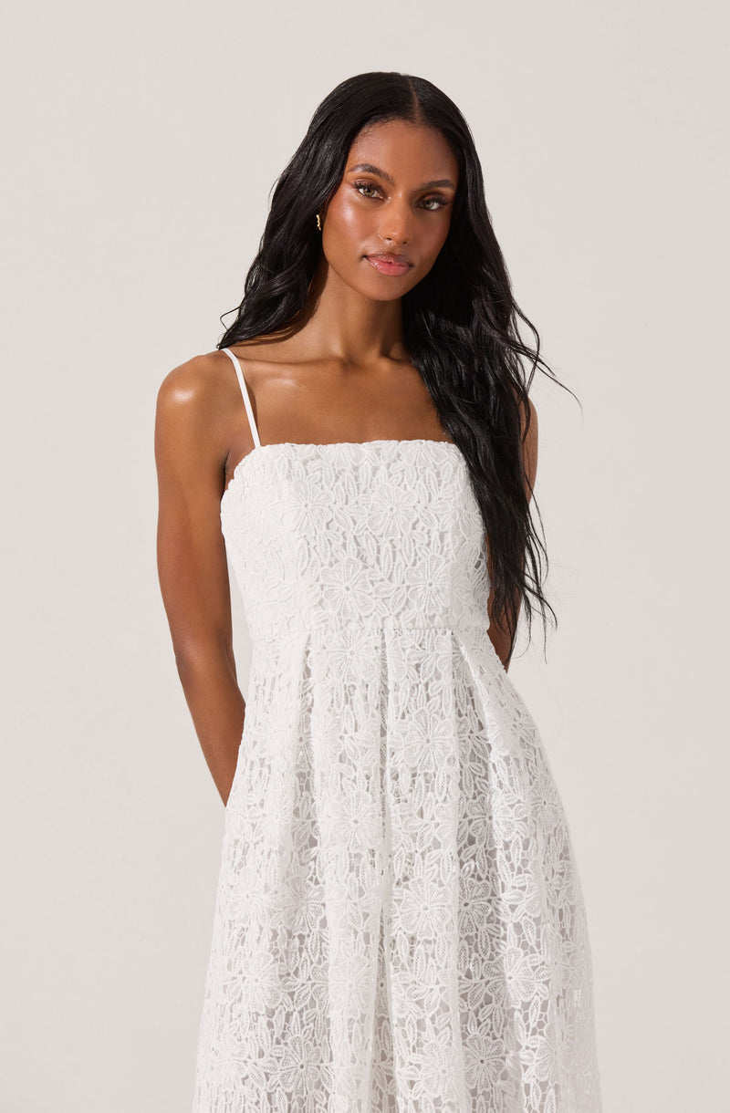 A woman is wearing a white sleeveless dress with thin spaghetti straps. The dress has a textured floral lace pattern covering the entire garment, creating a delicate and intricate appearance. The dress features an empire waistline with a gathered skirt that falls to the floor.