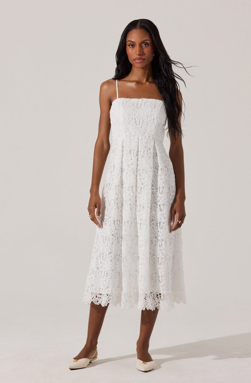 A woman is wearing a white, sleeveless, midi-length lace dress with thin spaghetti straps. The dress has a fitted bodice and a flared skirt with a scalloped lace hem. She pairs the dress with beige heels.