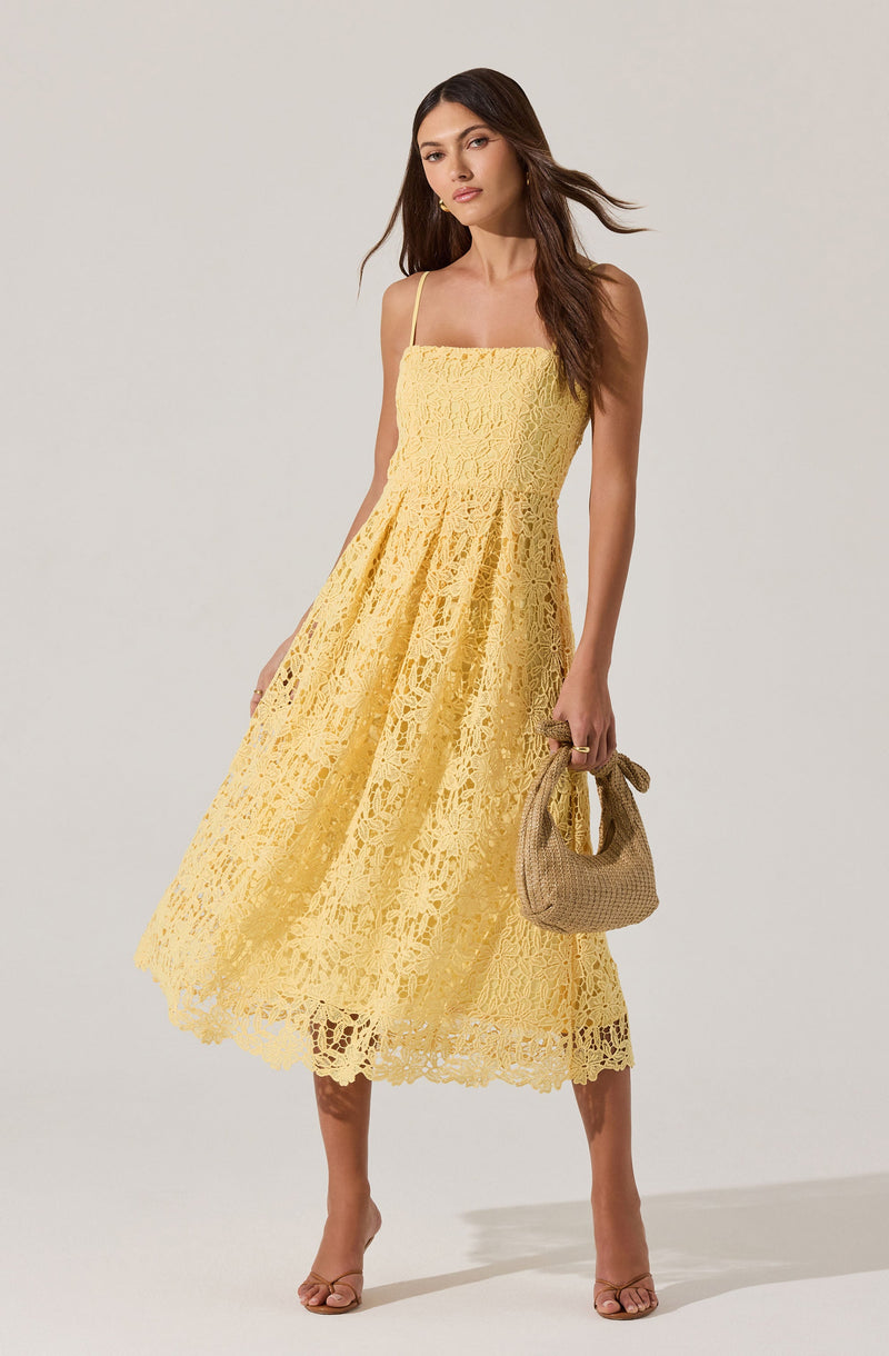 A woman is wearing a sleeveless, yellow, lace dress with thin spaghetti straps. The dress features a fitted bodice and a full, mid-length skirt with intricate floral lace patterns. She is holding a small, beige, woven handbag in her right hand, and she is wearing nude-colored, open-toe heels.