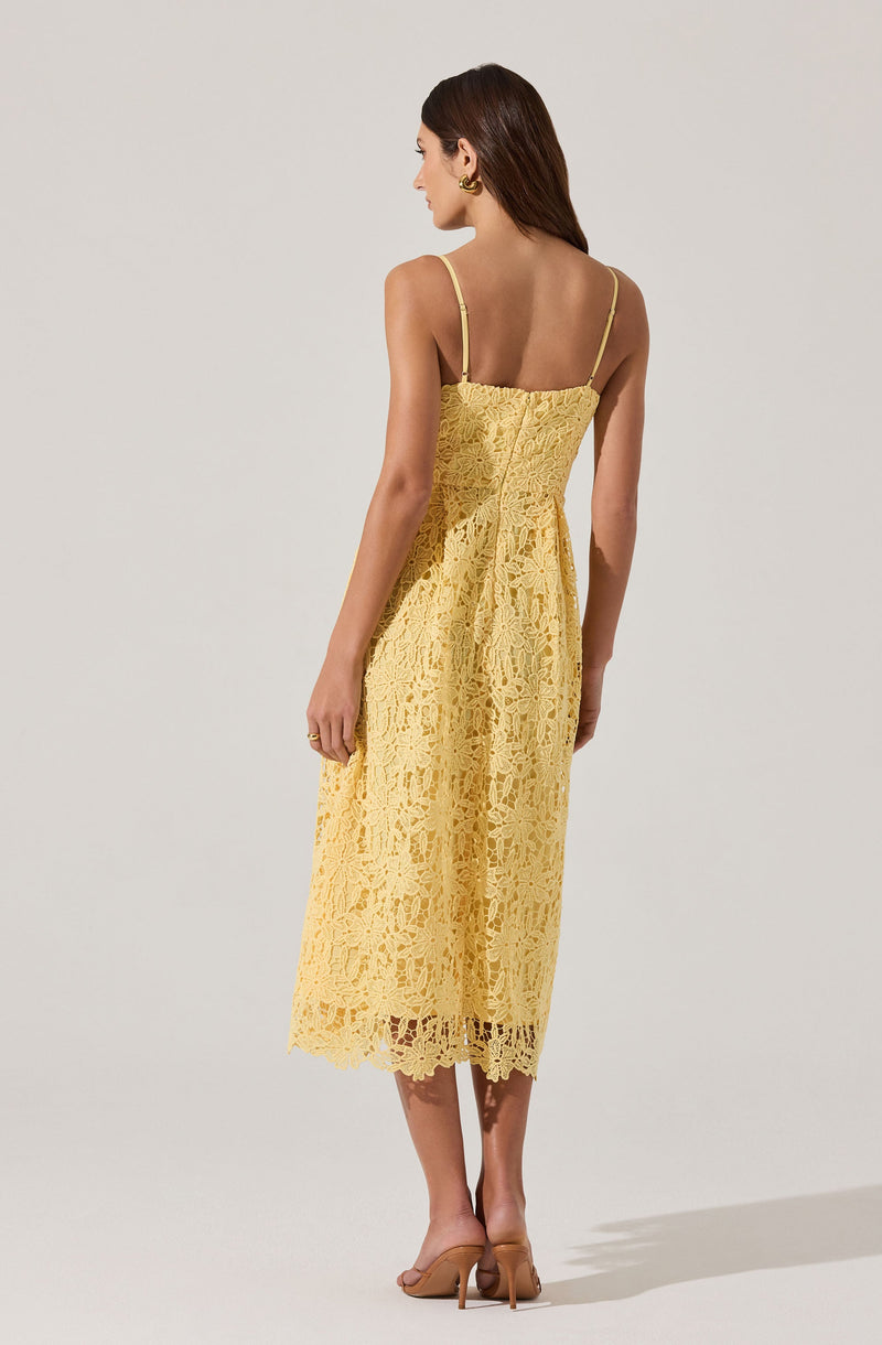 A woman is wearing a yellow lace midi dress with thin spaghetti straps. The dress features an intricate floral lace pattern and a fitted bodice that flows into a slightly flared skirt, ending below the calves. The lace has a scalloped hemline, adding a delicate touch to the overall look. She is also wearing nude high-heeled shoes and small earrings.