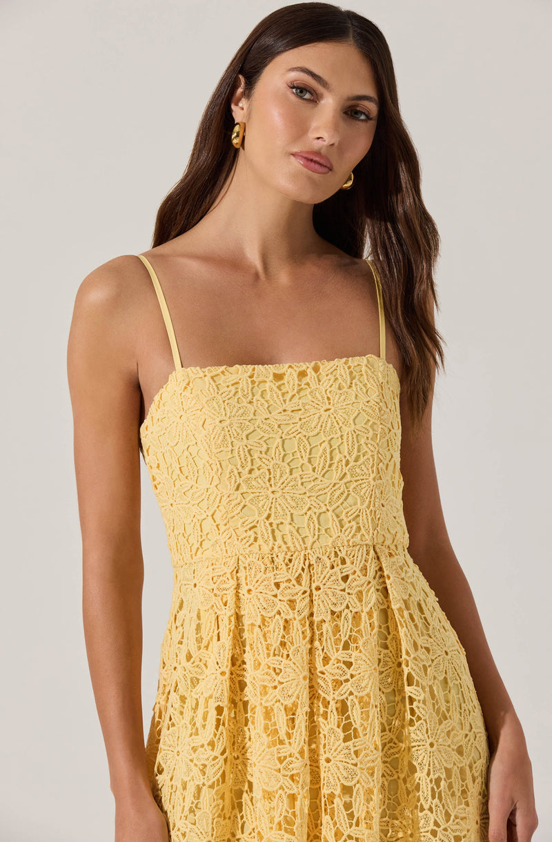 A woman is wearing a sleeveless yellow dress made of lace fabric with intricate floral patterns. The dress features thin spaghetti straps and a fitted bodice, flowing into a pleated skirt. She is accessorized with gold hoop earrings.
