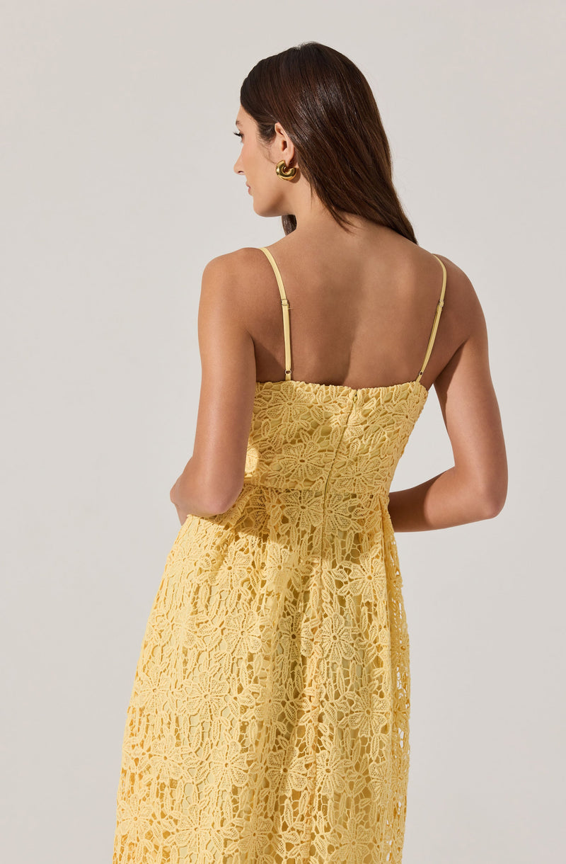 A woman is wearing a yellow dress with thin spaghetti straps. The dress features intricate floral lace patterns throughout, creating a textured and airy appearance. The back of the dress is fitted, and the lace fabric extends to the full length of the skirt. The woman is also wearing large, gold hoop earrings.