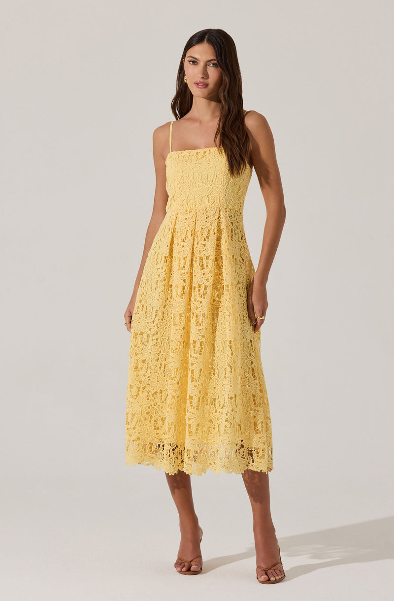 A woman is wearing a yellow lace dress with thin spaghetti straps. The dress has a fitted bodice and a full, midi-length skirt made of intricate lace fabric. The lace pattern is floral and open-work, creating a textured and airy look. She is also wearing neutral-colored open-toe heels.