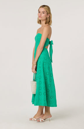 A woman is wearing a strapless, turquoise dress with eyelet embroidery. The dress has a fitted bodice and a high waist, with a matching sash tied in a bow on her left side. The skirt is mid-calf length, flowy, and features a circular eyelet pattern. She is also wearing white high-heeled sandals with thin straps.